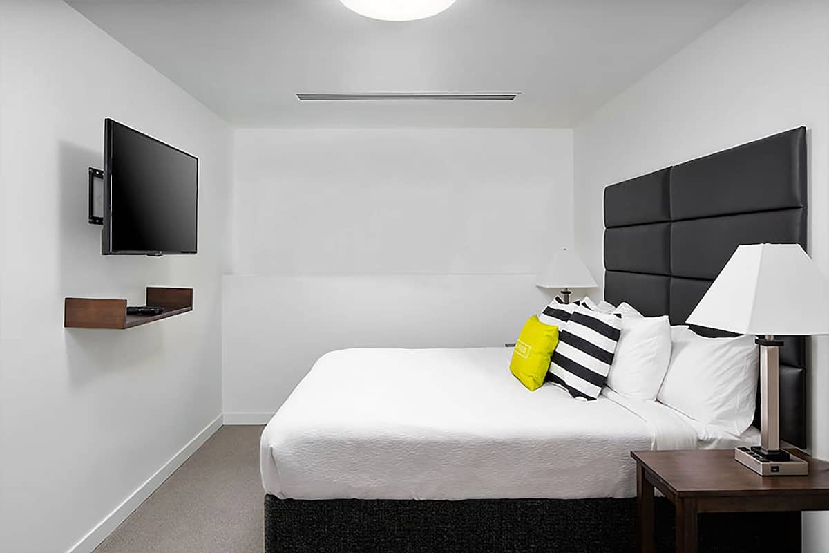 A modern hotel room with a large bed, a flat-screen TV mounted on the wall, a bedside table with a lamp, and a small shelf.