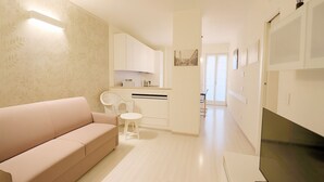 Una sala moderna con un sofá, una mesita y una silla. Se observa una zona de cocina con armarios y un ventanal.
