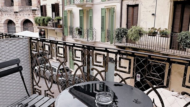 Un balcón con una mesa y sillas, una baranda de metal y edificios con contraventanas verdes al fondo.
