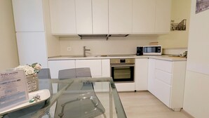 Una cocina compacta con armarios blancos, microondas, horno y una mesa de comedor de vidrio.