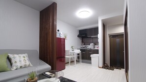 A compact living space with a kitchenette, a sofa with a green pillow, a glass-top table with a remote control, and a small plant. - Ko's Apartment, Tatsumi West Station (Osaka)