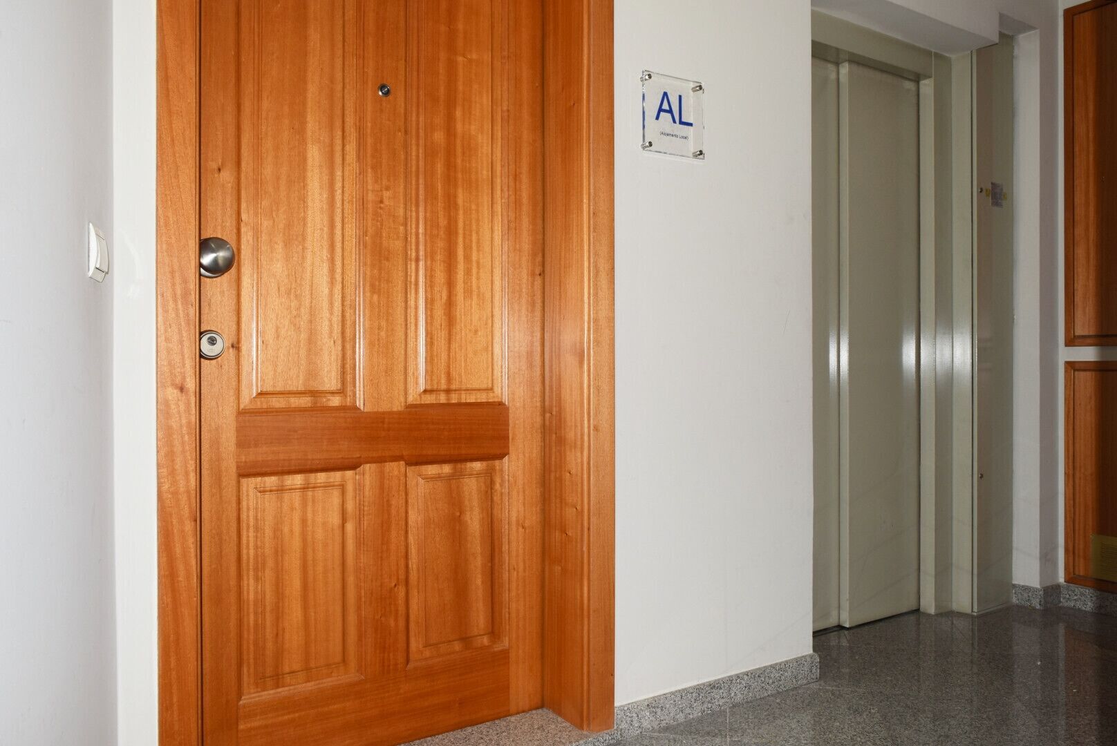 A wooden door with a metal handle, a white wall with a sign reading "AL", and a hallway with a tiled floor.