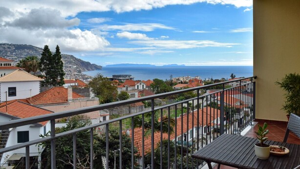 Un balcon offrant une vue sur une ville côtière, avec des maisons aux toits recouverts de tuiles rouges et une vue lointaine sur la mer.