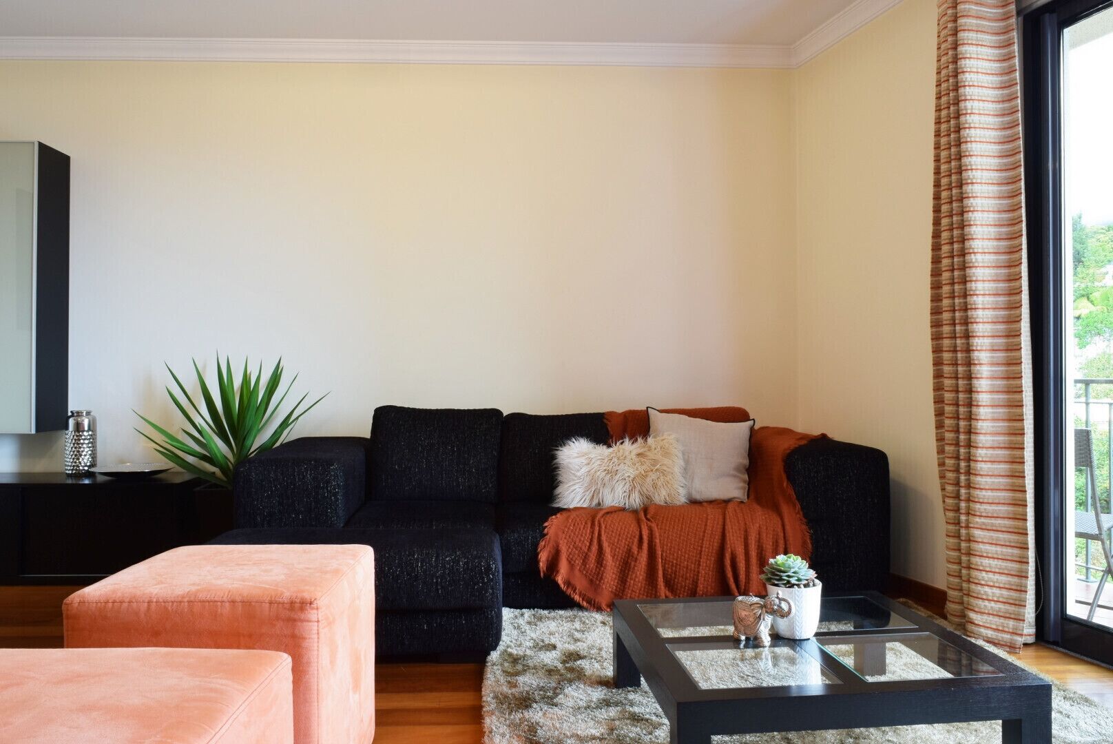 A living room with a black sofa, a peach-colored ottoman, a glass coffee table with a decorative item, and a large plant.