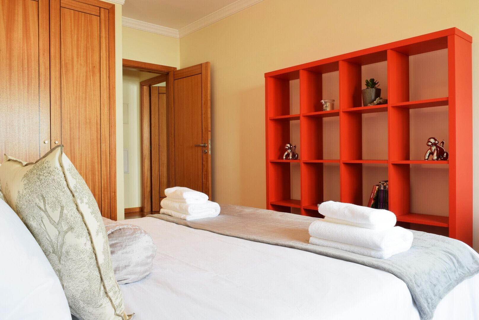 A hotel room with a bed, a wooden wardrobe, and a red shelving unit with decorative items.