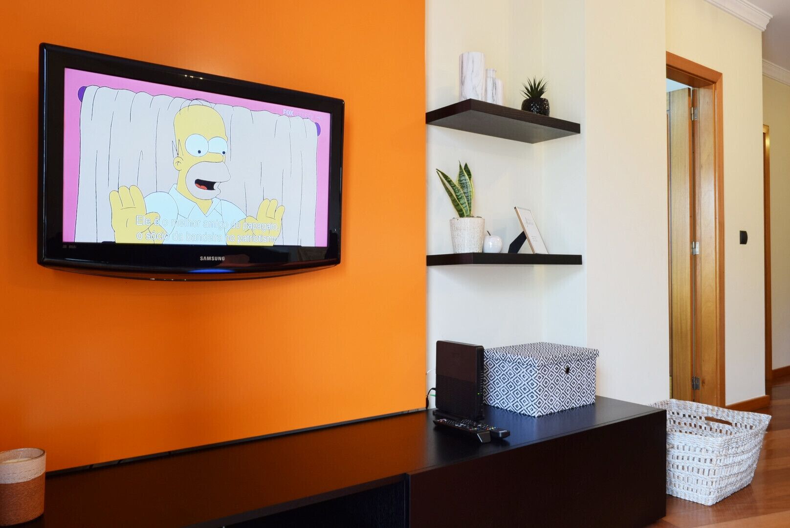 A television mounted on an orange wall, a black console table, and two black shelves with decorative items.