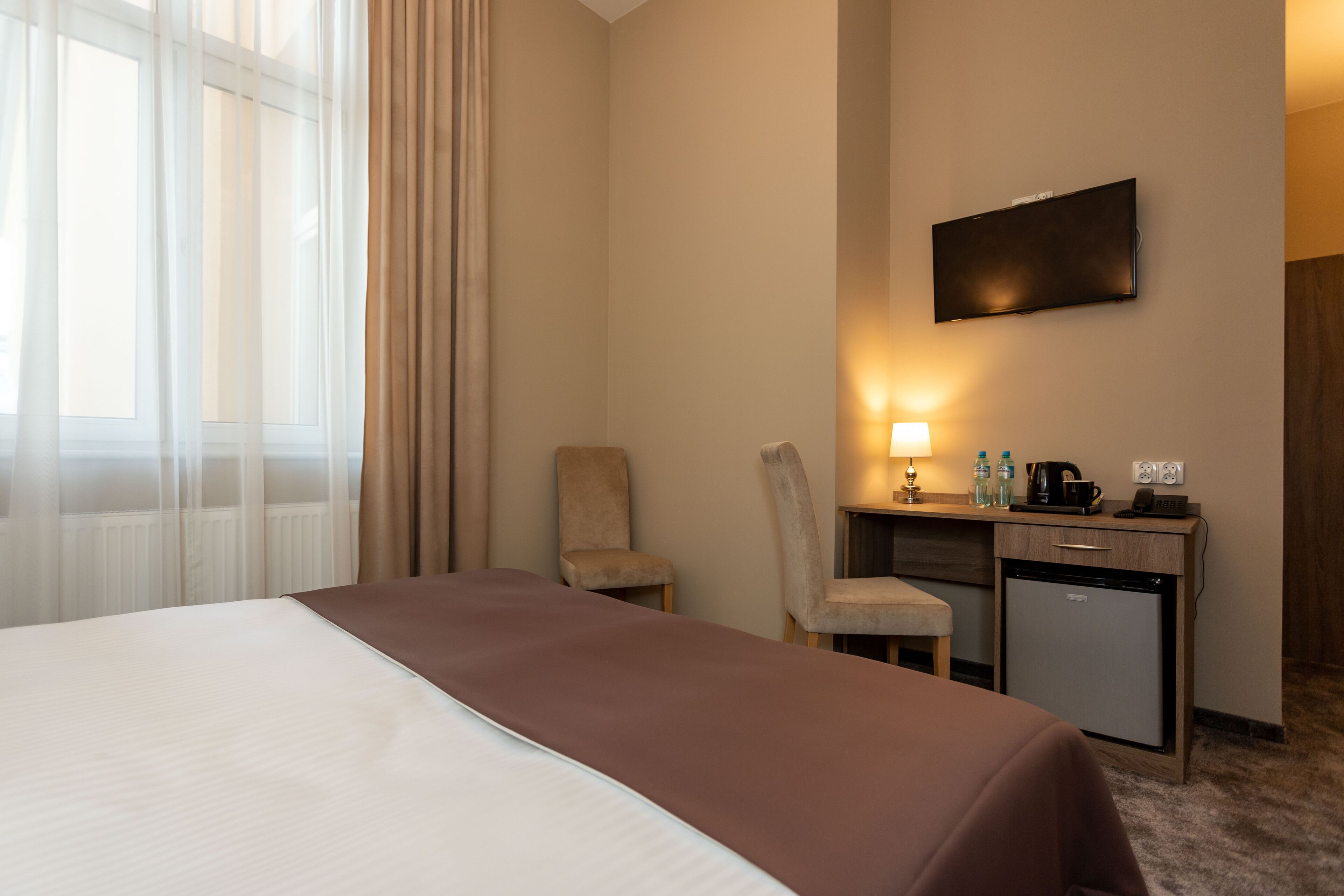 Una habitación de hotel con una cama, un escritorio con lámpara, una silla y un refrigerador pequeño.