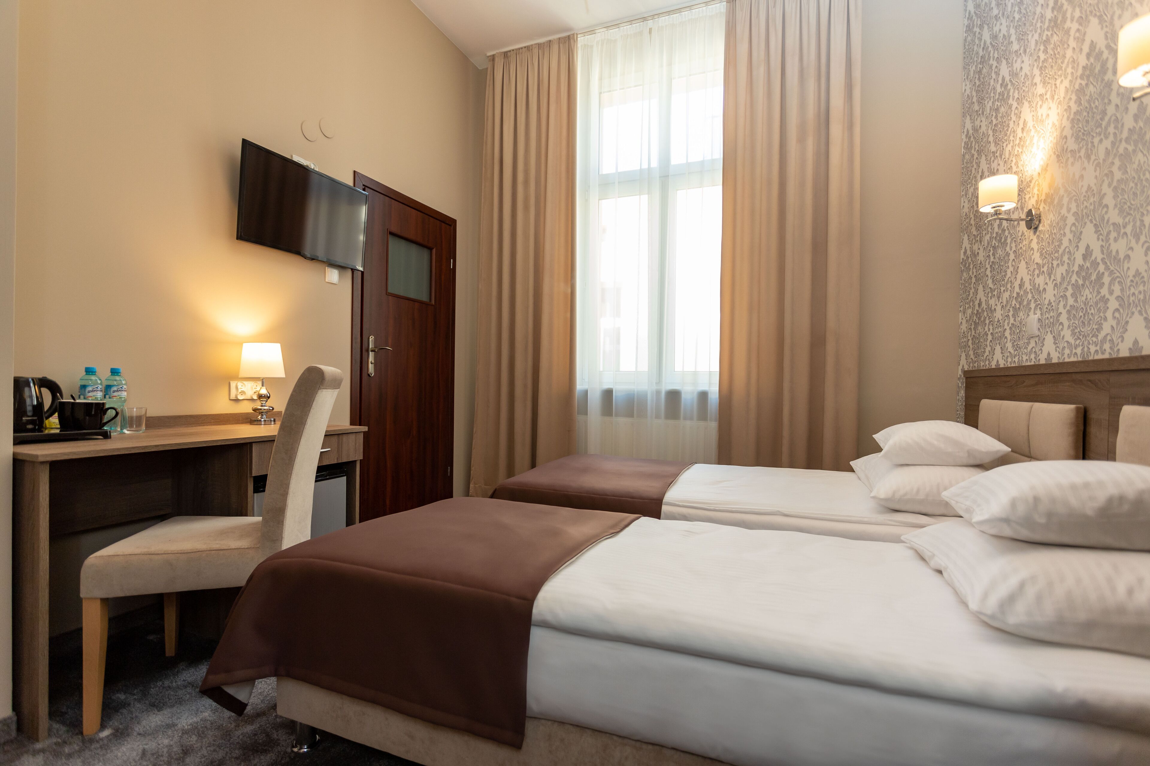 Una habitación de hotel con dos camas, un escritorio con silla, televisor y ventana con cortinas.