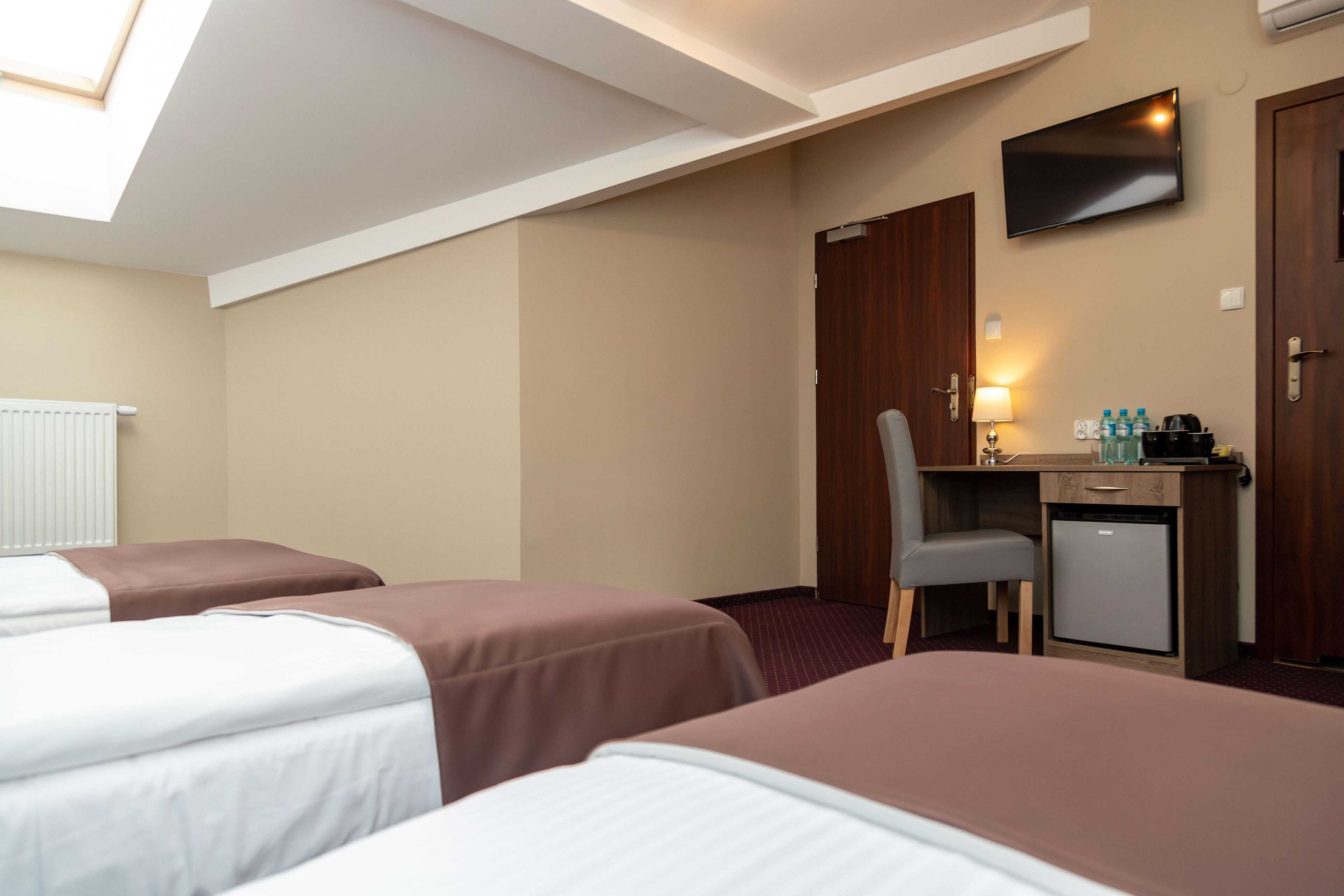 A hotel room with two beds, a desk with a chair, a small refrigerator, a TV mounted on the wall, and a window with blinds.