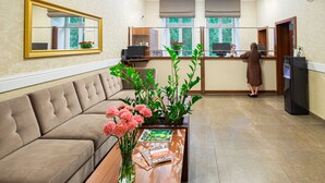 Zona de recepción de un hotel con un sofá, una mesa de centro con flores, un espejo y un mostrador de recepción con una computadora.