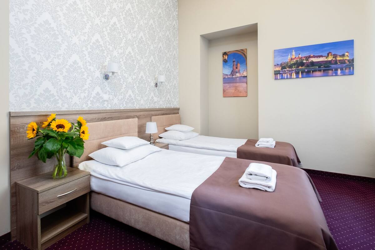 Habitación de hotel con dos camas, una mesita de noche, un florero con girasoles y un cuadro de un paisaje urbano.