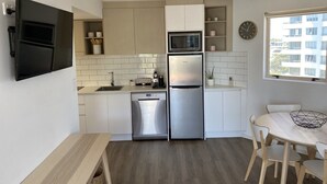 A compact kitchen with a built-in microwave, stainless steel refrigerator, and dishwasher. There is a dining area with a table and chairs, and a window providing a view of the building outside. - Mooloolaba Beachfront Apartment (Mooloolaba)