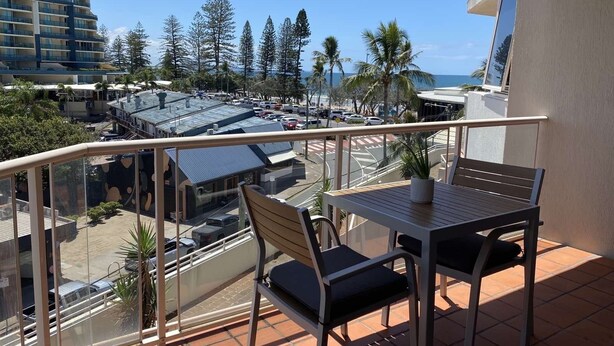A balcony with a table and chairs, overlooking a parking area and buildings with a clear view of the ocean. - Mooloolaba Beachfront Apartment (Mooloolaba)