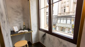 Una habitación pequeña con una ventana, un taburete de madera, un estante con un libro y una botella, y una pared de mármol.