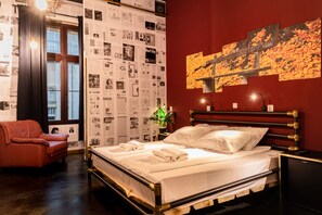 Ein ordentlich bezogenes Bett mit weißen Leinentüchern und Handtüchern, ein roter Ledersessel, eine Wand mit einem großen abstrakten Gemälde und eine mit Postern beklebte Wand.