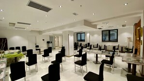 Una amplia sala de conferencias con varias mesas y sillas, paredes blancas e iluminación en el techo.