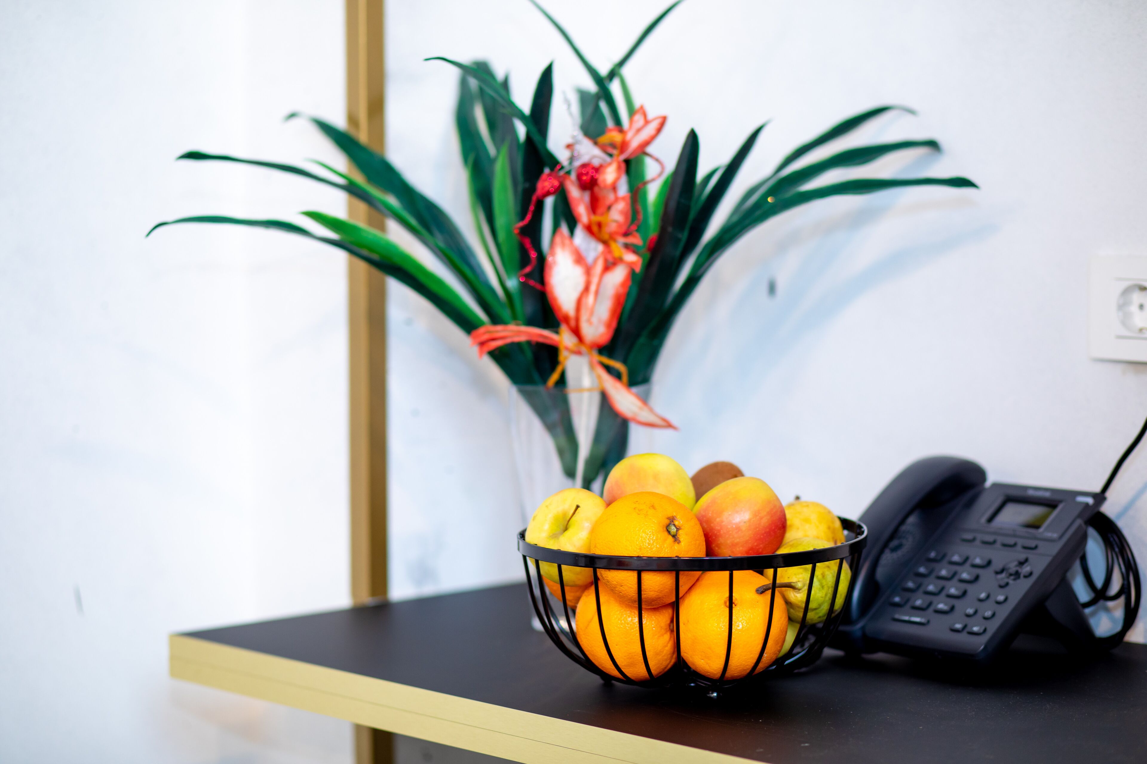 Un escritorio con un frutero que contiene naranjas y manzanas, un teléfono y una planta en maceta.