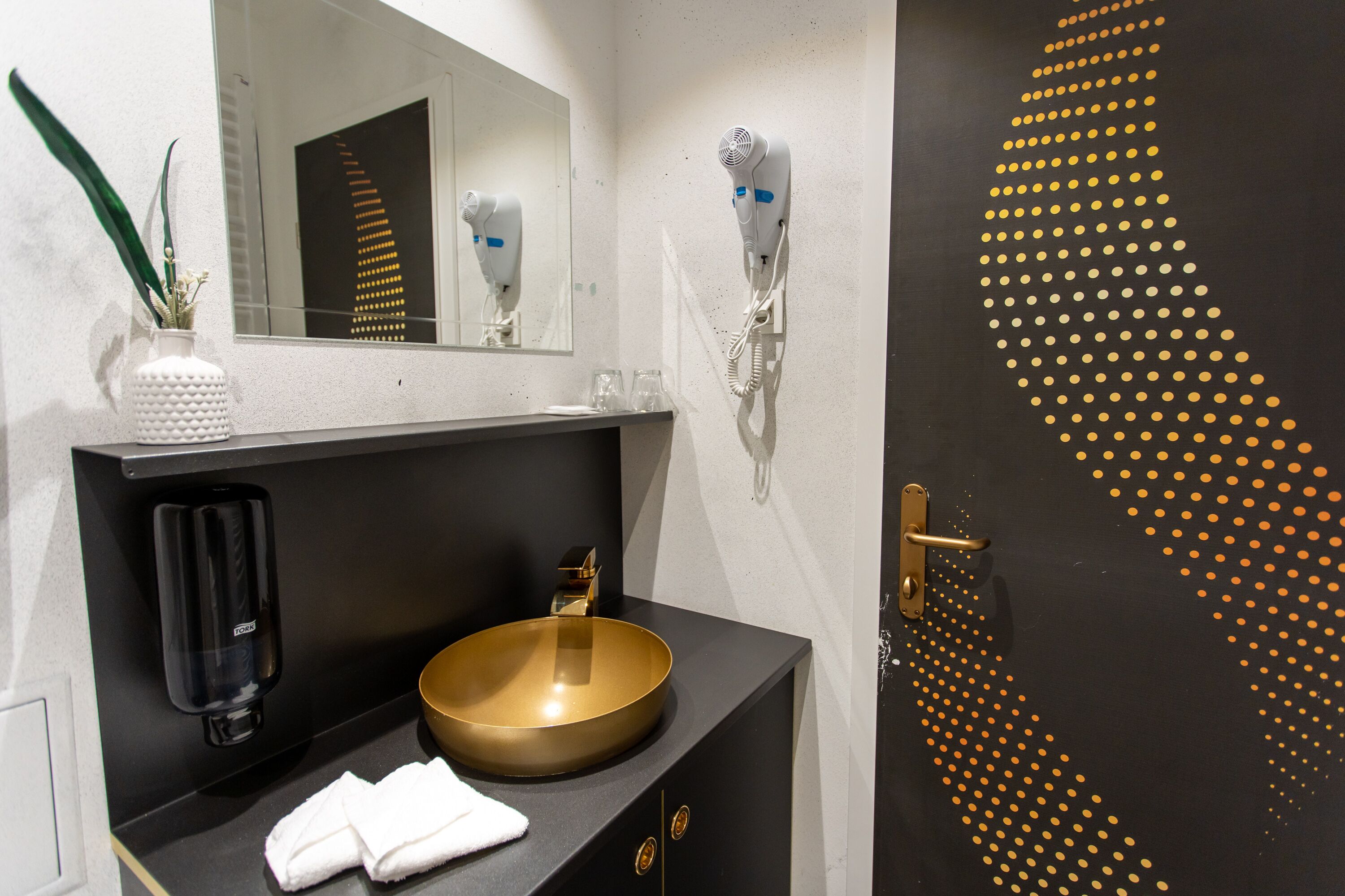 Ein Badezimmer mit einem runden, goldenen Waschbecken, einem Handtuchhalter, einem Spiegel, einem Föhn und einem dekorativen Wandmuster.