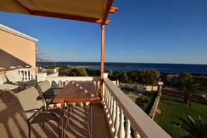 A balcony with a table and chairs, overlooking a coastal landscape with a clear sky.
