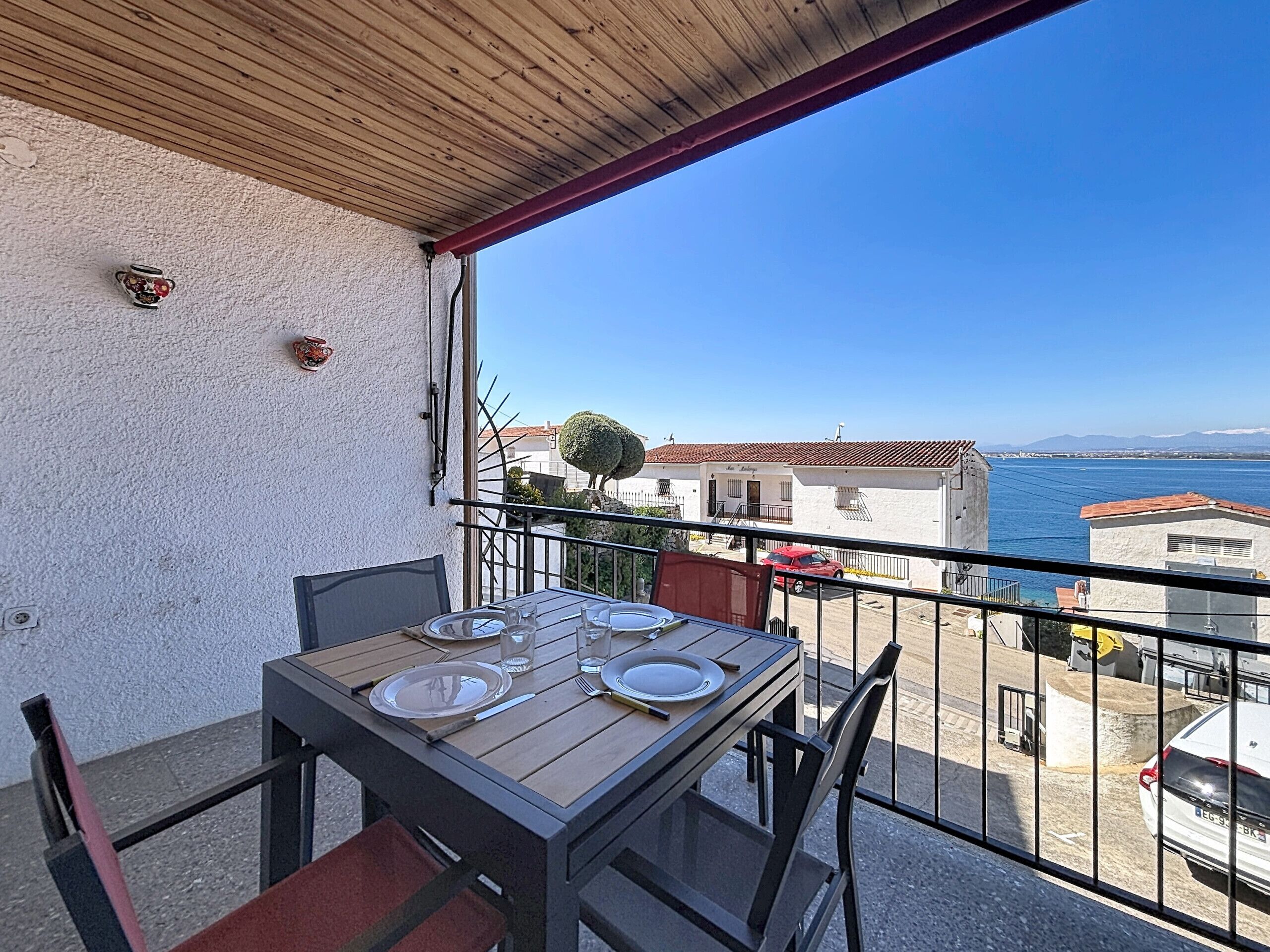 A balcony with a table set for two, overlooking a coastal area with buildings and a clear sky.