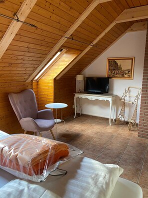 A cozy attic room with a sloped ceiling, a television, a white desk, a cushioned chair, and a bed with a blanket.