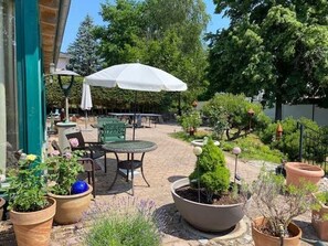 An outdoor patio with tables and chairs, surrounded by potted plants and a white umbrella.