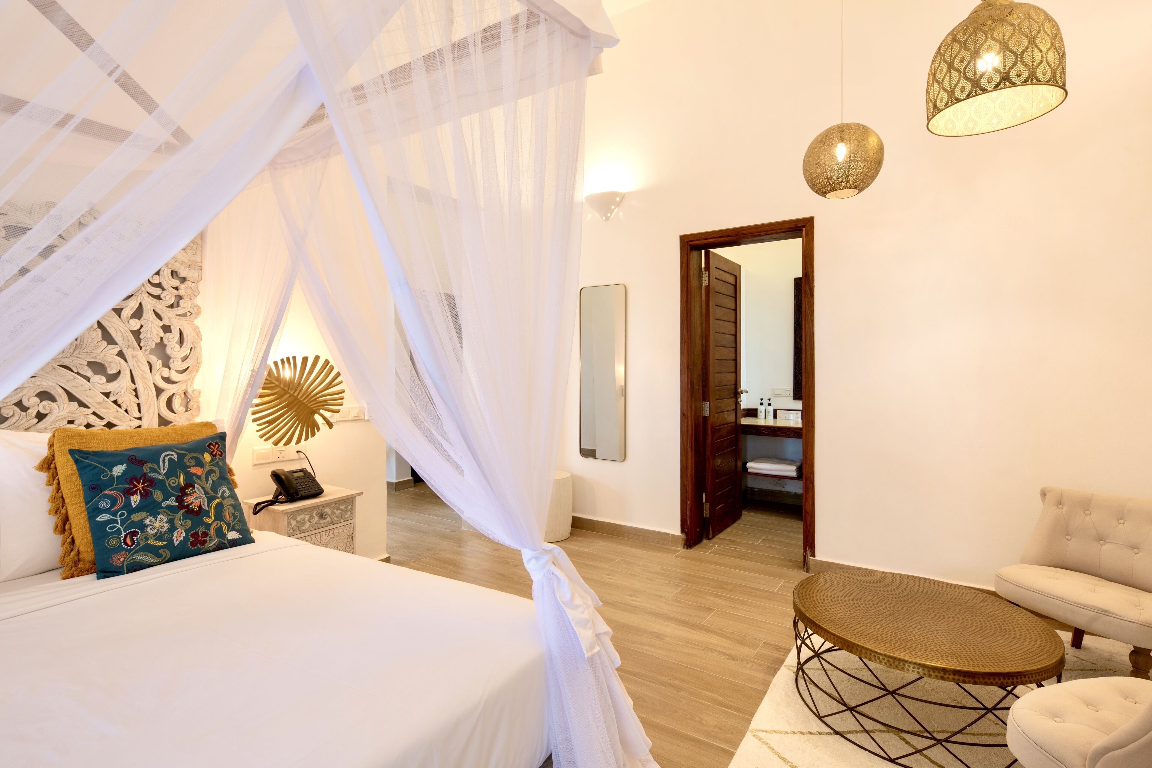 A bedroom with a bed, a bedside table, a lamp, a mirror, and a door leading to another room.