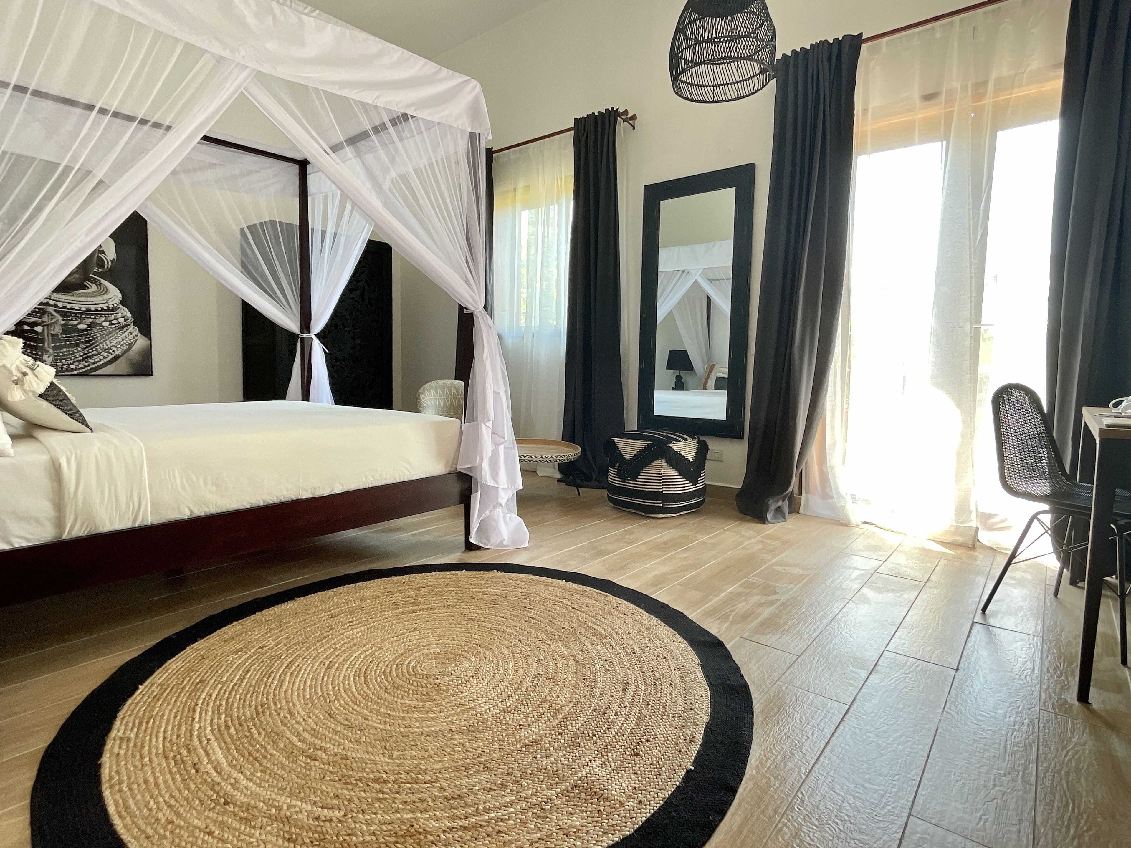 A bedroom with a bed, a round woven rug, a bedside table, a chair, a mirror, and a window with curtains.