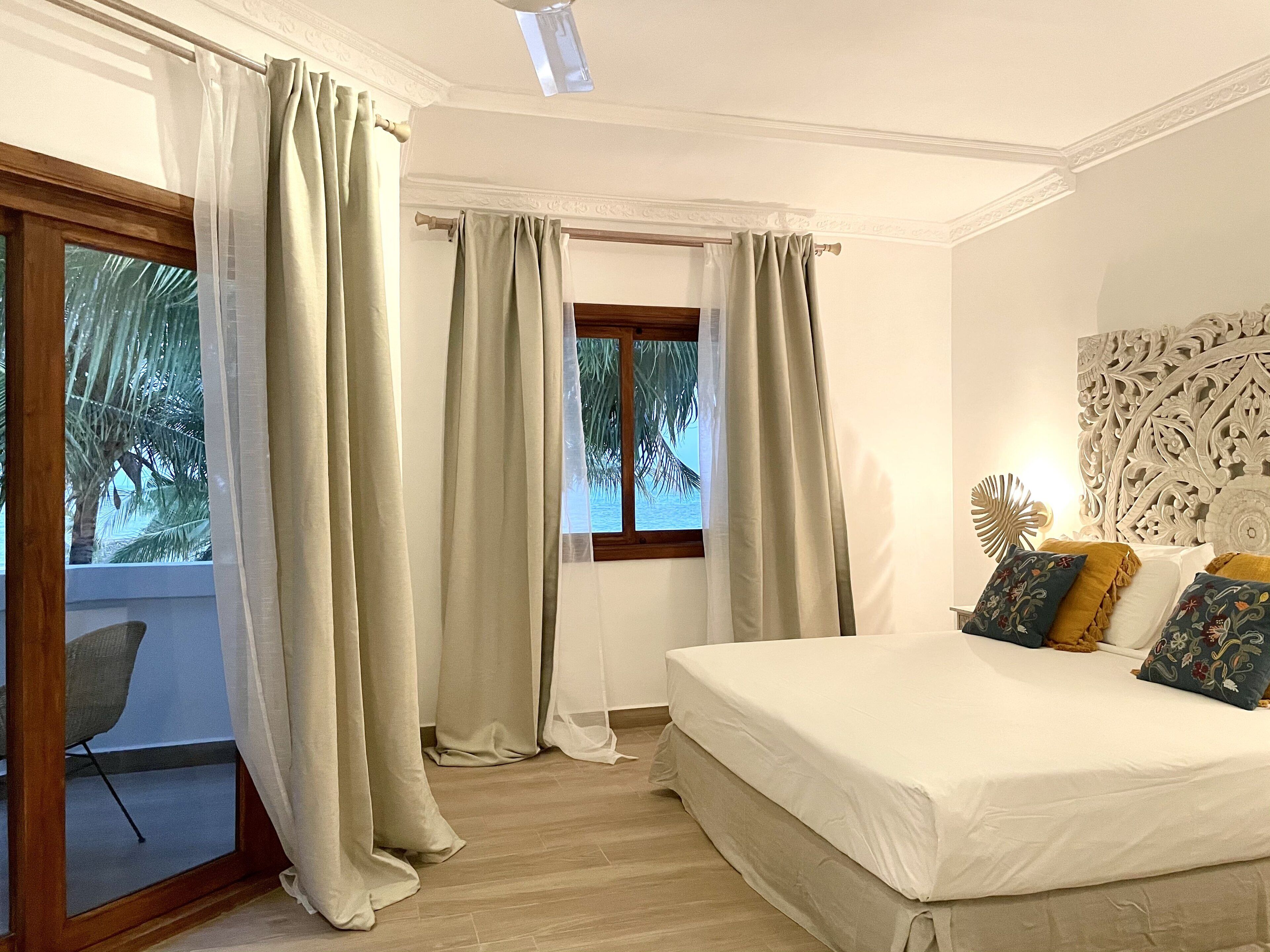 A bedroom with a bed, curtains, a window with a view of palm trees, and a decorative wall piece.
