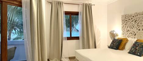 A bedroom with a bed, curtains, a window with a view of palm trees, and a decorative wall piece.
