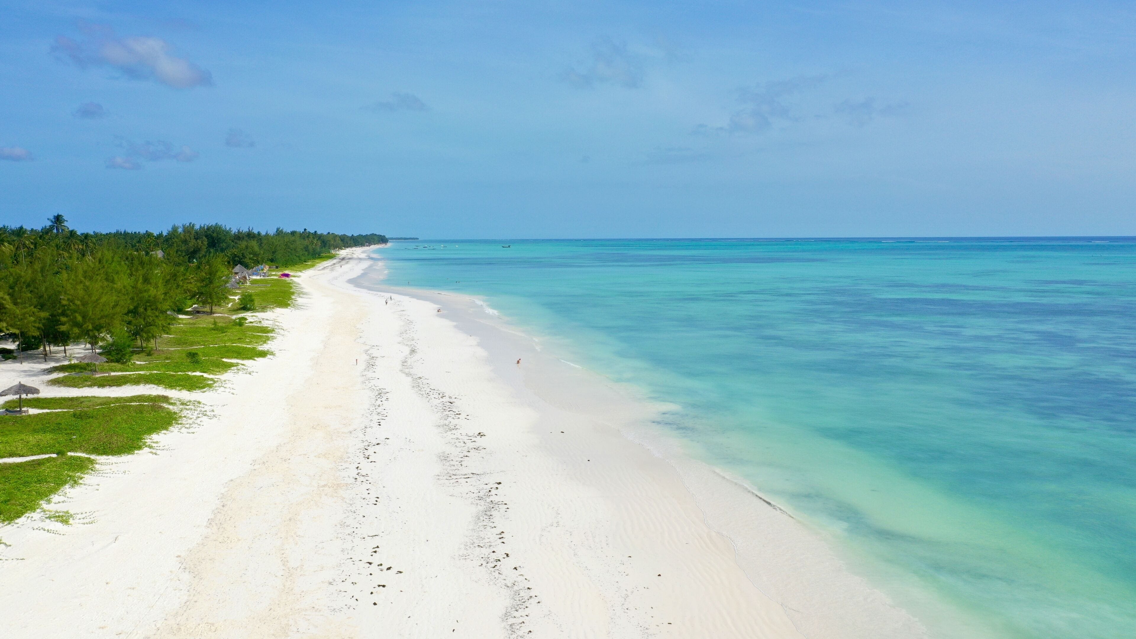A wide sandy beach with clear turquoise water, lined with green vegetation and a few structures on the left.