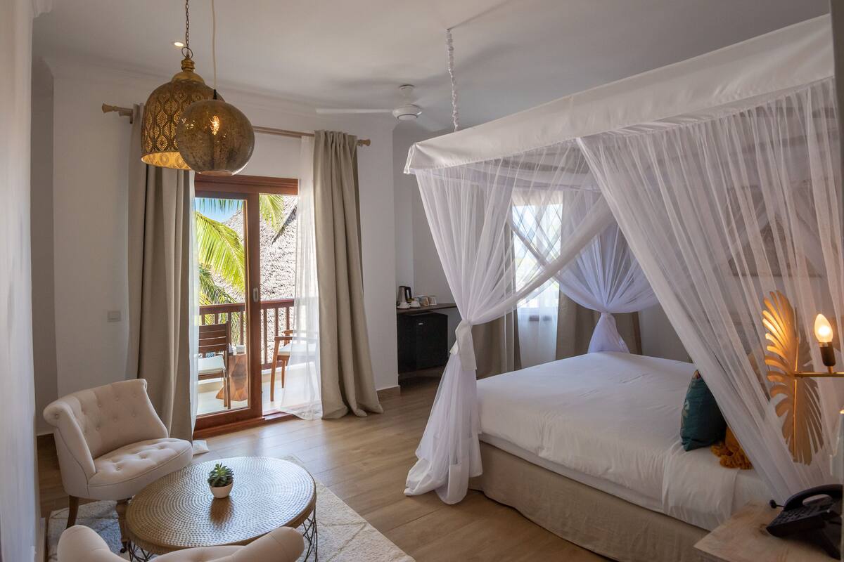 A bedroom with a canopy bed, a round table with a plant, a chair, and a desk with a lamp.