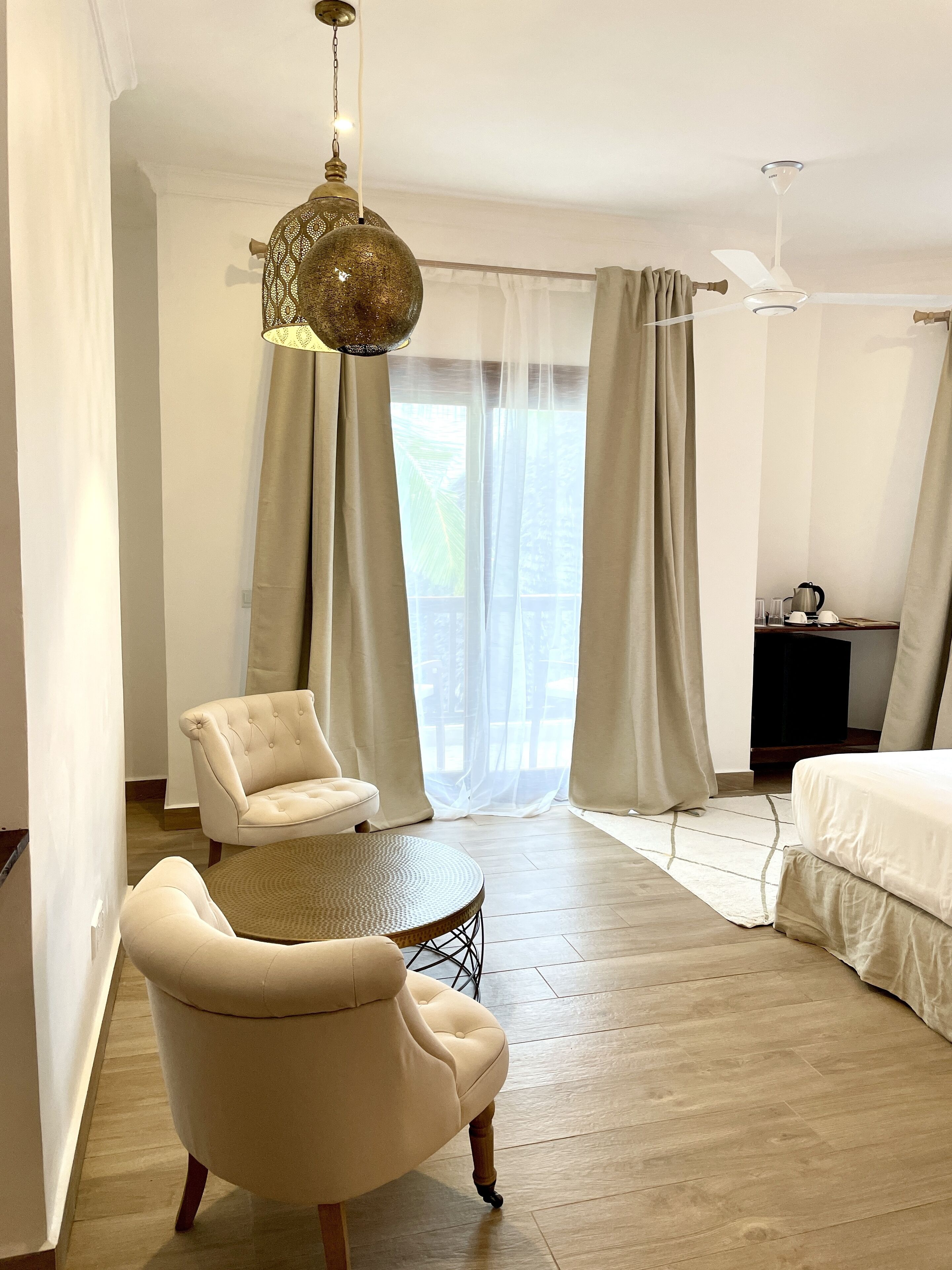 A hotel room with a bed, a round table, two armchairs, a ceiling fan, and a large window with sheer curtains.