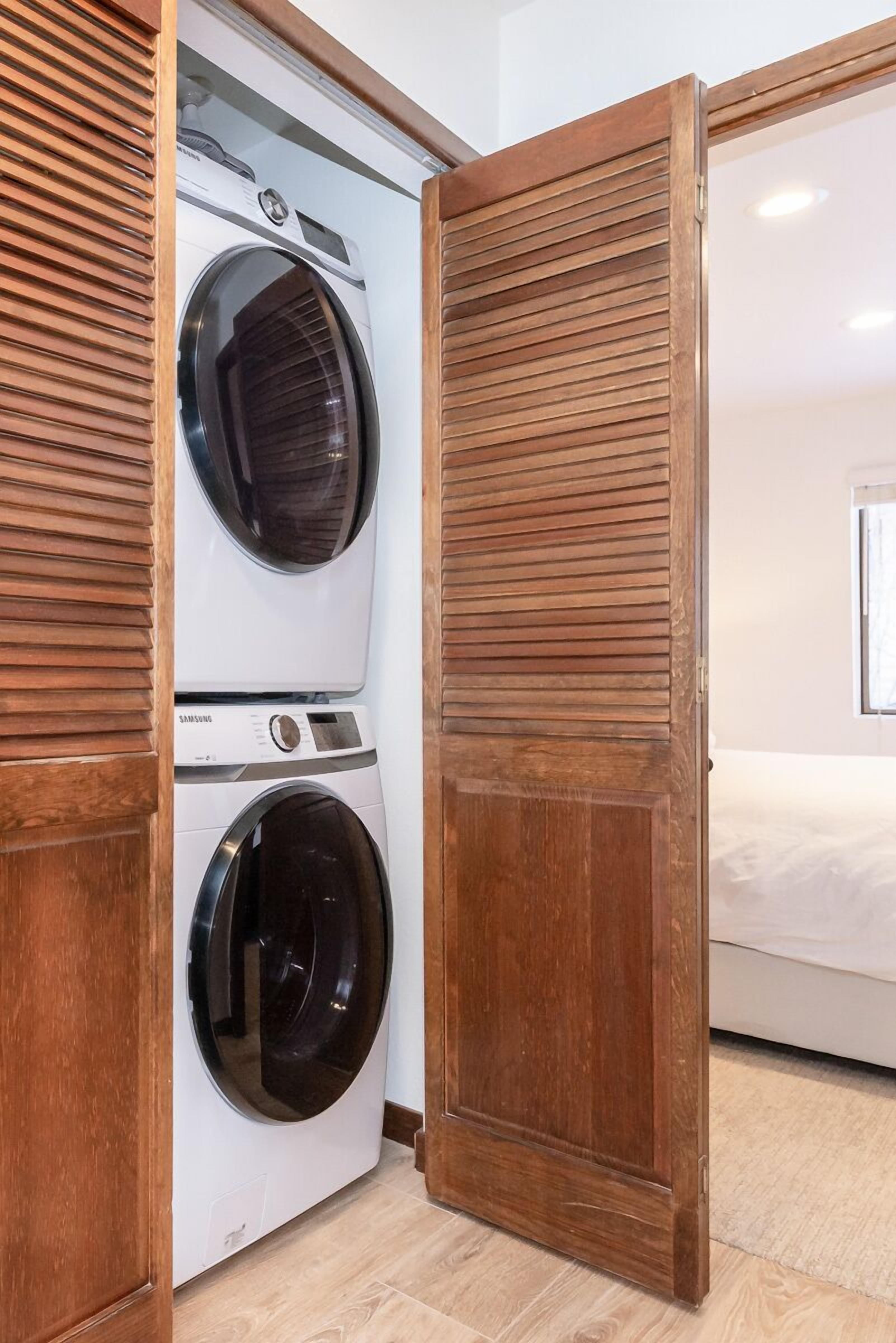 Una lavandería con dos lavadoras apiladas, una puerta de madera y una cama en la habitación contigua.