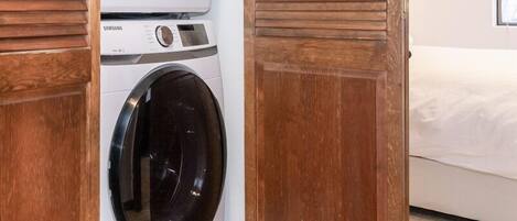 Una lavandería con dos lavadoras apiladas, una puerta de madera y una cama en la habitación contigua.