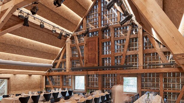 Un comedor rústico con vigas de madera y ventanas amplias, mesas dispuestas para una comida.