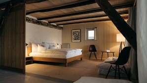 Un dormitorio con techo de madera, una cama, una silla, una mesita y una ventana con cortinas.