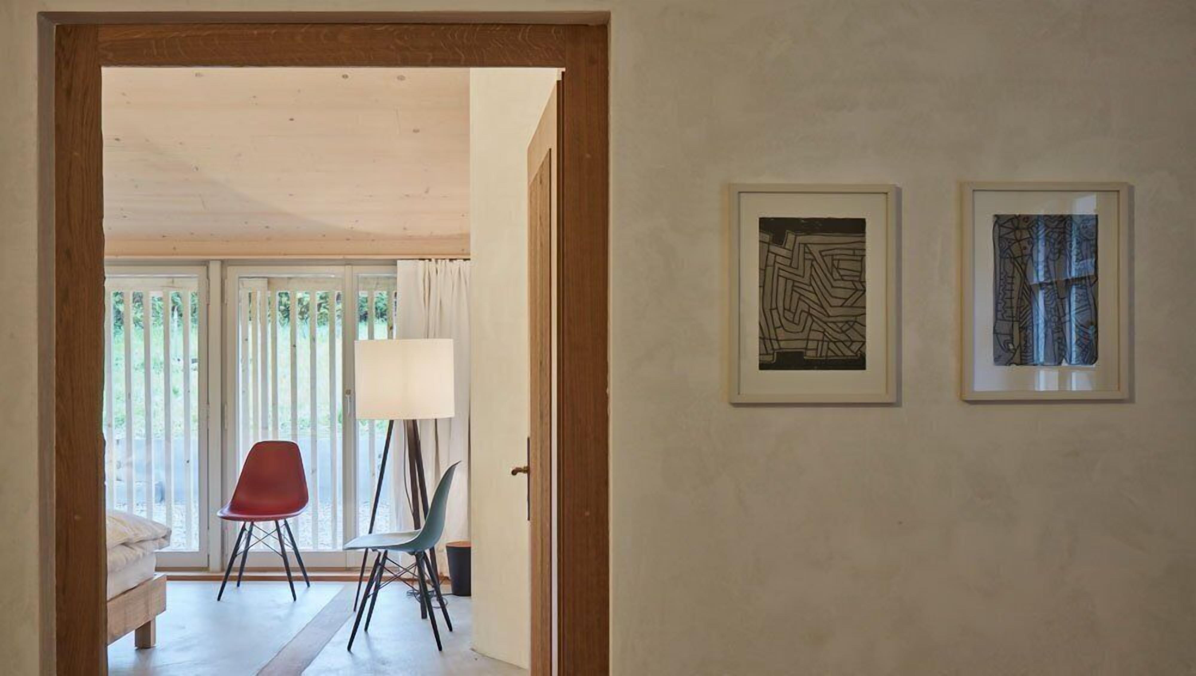 Un pasillo con un marco de puerta de madera, una silla roja, una lámpara de pie y dos obras de arte abstracto enmarcadas en la pared.