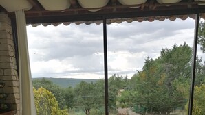Una habitación con un amplio ventanal que ofrece vistas a una zona boscosa. Hay un sofá beige con cojines y suelo de piedra.