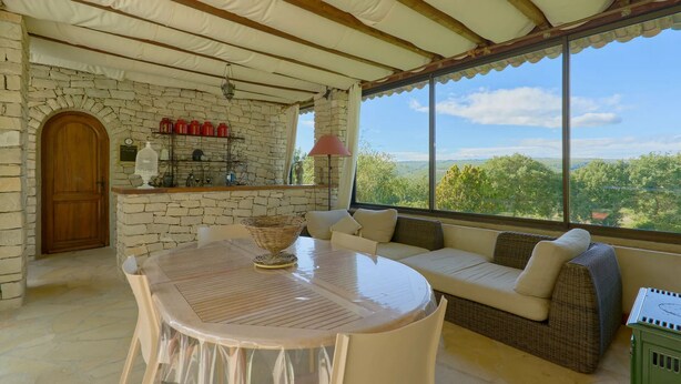 Una habitación acogedora con pared de piedra, puerta de madera, una mesa redonda con una cesta, sofá con cojines y vistas del paisaje a través de grandes ventanales.