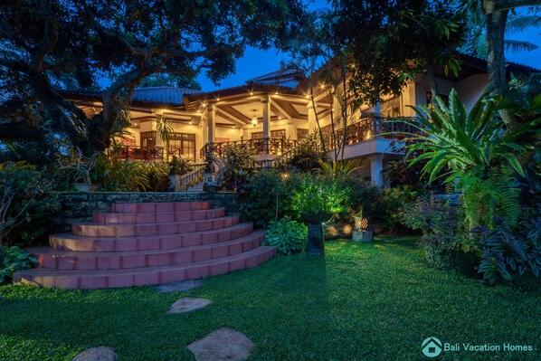 A large, well-lit house with a covered porch, surrounded by lush greenery and a stone staircase leading to the entrance.