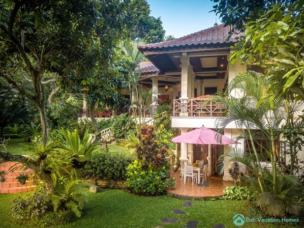 A two-story building with a balcony, surrounded by lush greenery and a patio area with outdoor furniture.