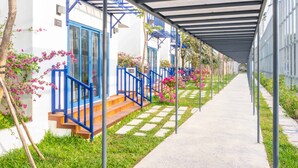 Une allée couverte avec des garde-corps bleus, des marches menant à un bâtiment aux fenêtres encadrées de bleu, et des plantes à fleurs roses le long du chemin.