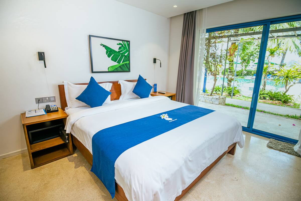 A hotel room with a large bed, a nightstand, a desk, and a window with a view of greenery.