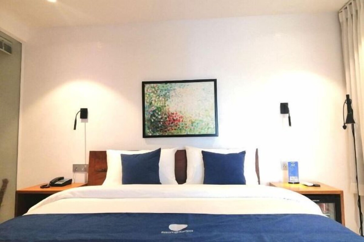 A hotel room with a neatly made bed, a painting on the wall, and a nightstand with a phone and a lamp.