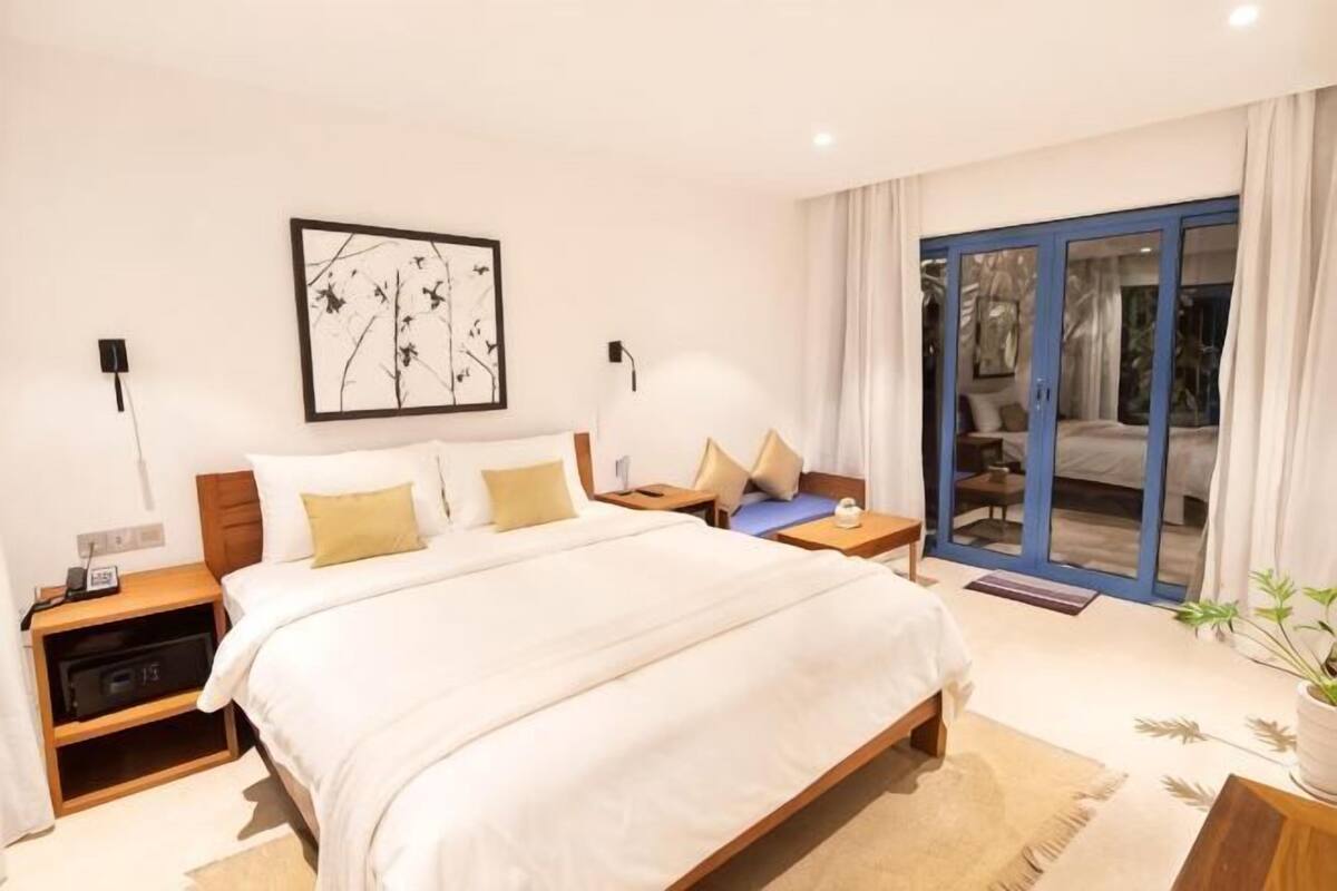 A bedroom with a bed, bedside tables, a nightstand, a wall-mounted lamp, a window with curtains, and a sliding glass door leading to a balcony.