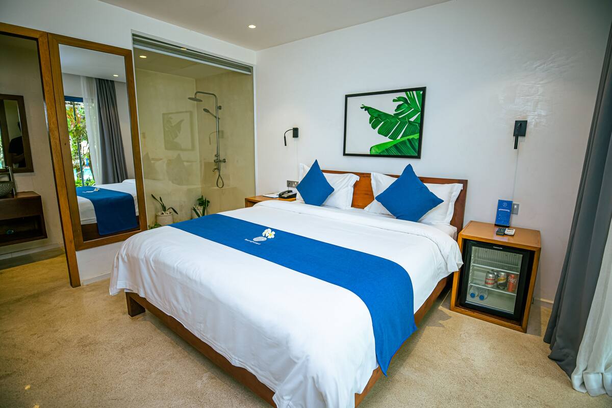 A hotel room with a large bed, a glass shower enclosure, a small refrigerator, and a framed plant on the wall.