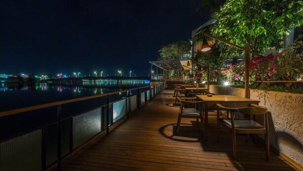 Une terrasse en bois, avec des tables et des chaises, donnant sur une étendue d’eau la nuit.