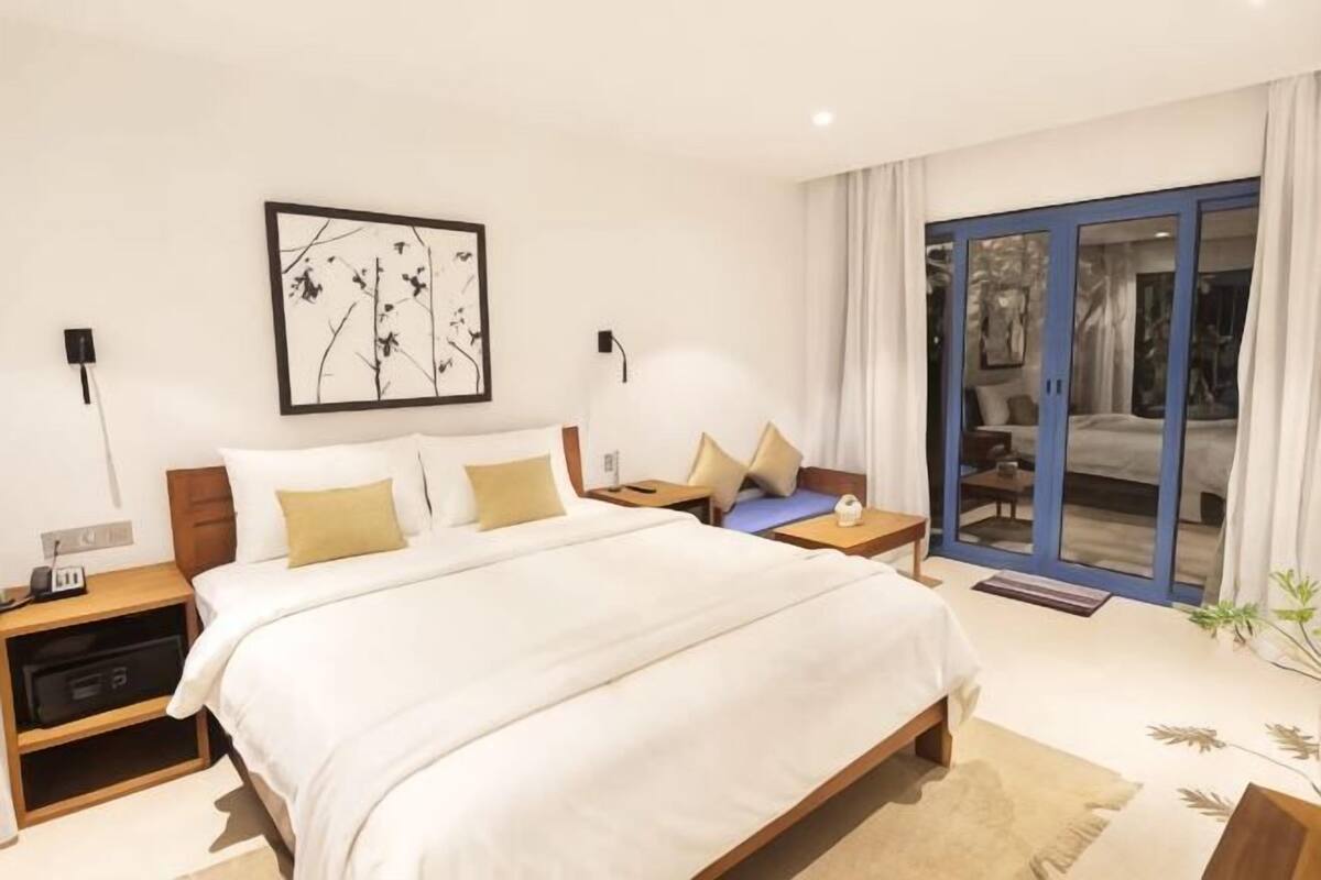 A modern hotel room with a large bed, bedside tables, a nightstand, a wall-mounted artwork, and a sliding glass door leading to a balcony.