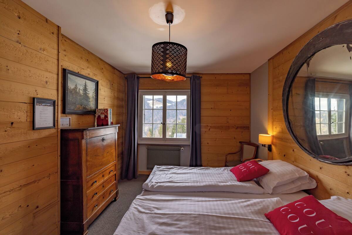 Een kamer met houten lambrisering, een bed, een dressoir, een raam met uitzicht en een hanglamp.